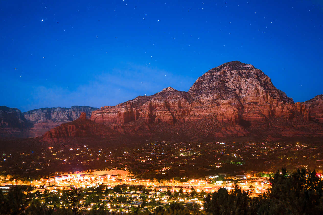 Nighttime view of Sedona with twinkling stars in the sky and city lights illuminating the valley below, framed by silhouettes of mountains